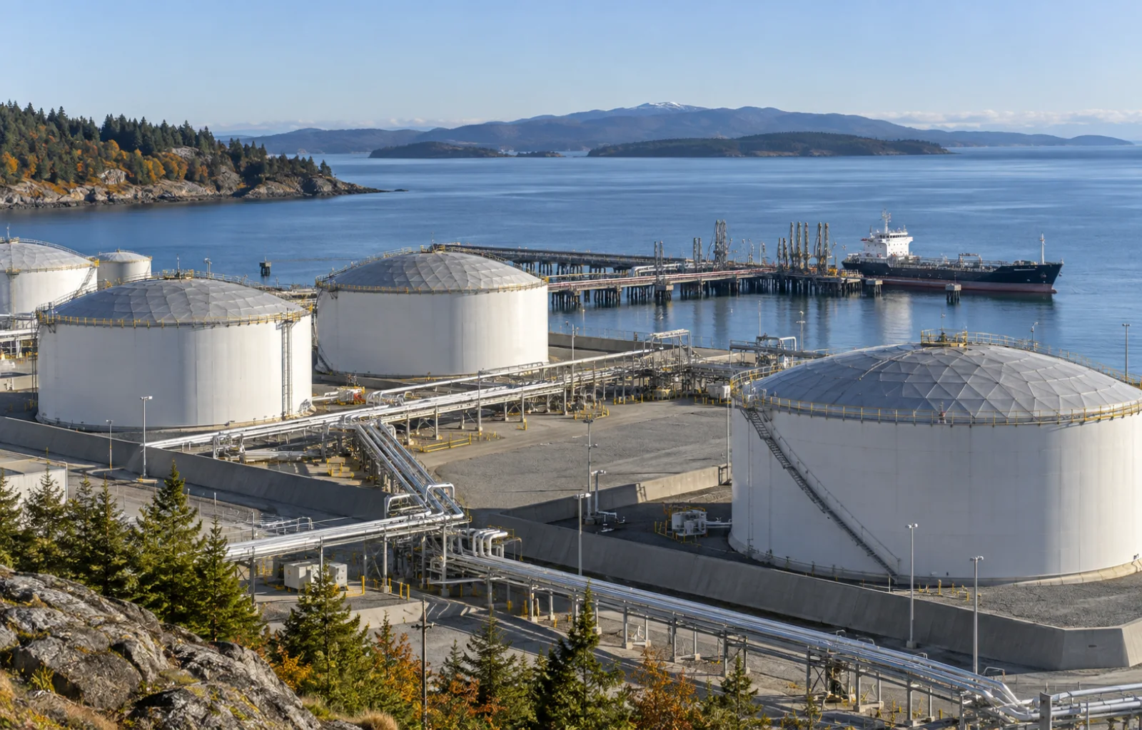 Fuel storage tanks and pipe racks stand beside a marine product-tanker berth at a quiet coastal distribution terminal.